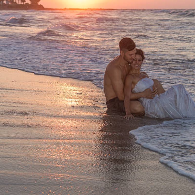 Just married young couple at the beach , in a late summer hazy day at dusk, wearing  a wedding dress and shorts, enjoying walking barefoot, getting wet, teasing and kissing one another.