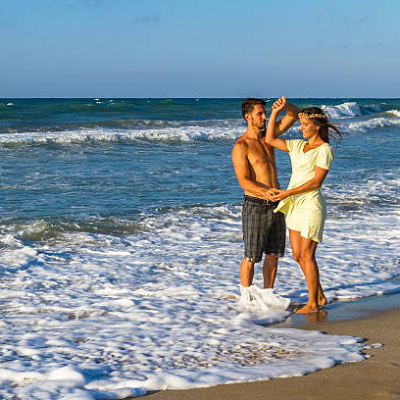 Happy attractive young couple in beachwear, enjoying a summer dusk at the beach, having fun walking barefoot, getting wet, kissing and teasing one another.